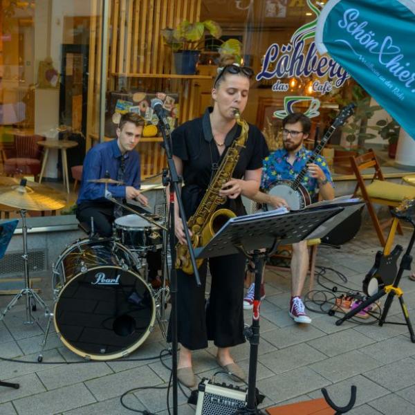 Drei Jazzmusiker*innen sind dargestellt, die vor einem Café-Schaufenster und neben einer hellblauen Schön hier!-Fahne gemeinsam auf einem Banjo, Saxofon und Schlagzeug musizieren | Foto: Walter Schernstein