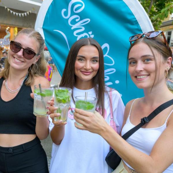 Drei junge Frauen halten gut gelaunt je einen Cocktail in die Kamera und stehen vor einer hellblauen Schön hier!-Fahne | Foto: Walter Schernstein