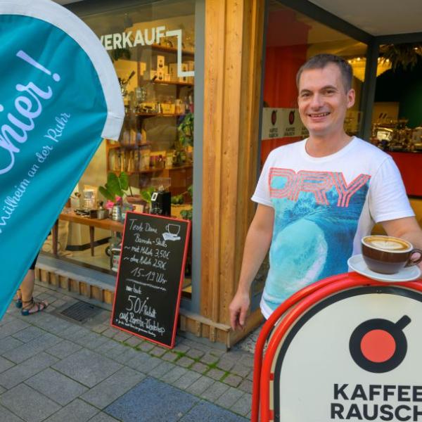 Ein junger Mann steht angelehnt an ein Werbeschild vor einem Geschäfts und hält eine Tasse Cappuccino in der Hand. Angeschnitten im Bild ist die hellblaue Schön hier!-Fahne zu sehen | Foto: Walter Schernstein