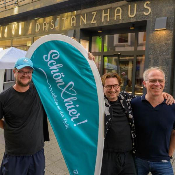 Drei Männer stehen vor einer Ladenzeile und halten die hellblaue Schön hier!-Fahne in der Hand | Foto: Walter Schernstein