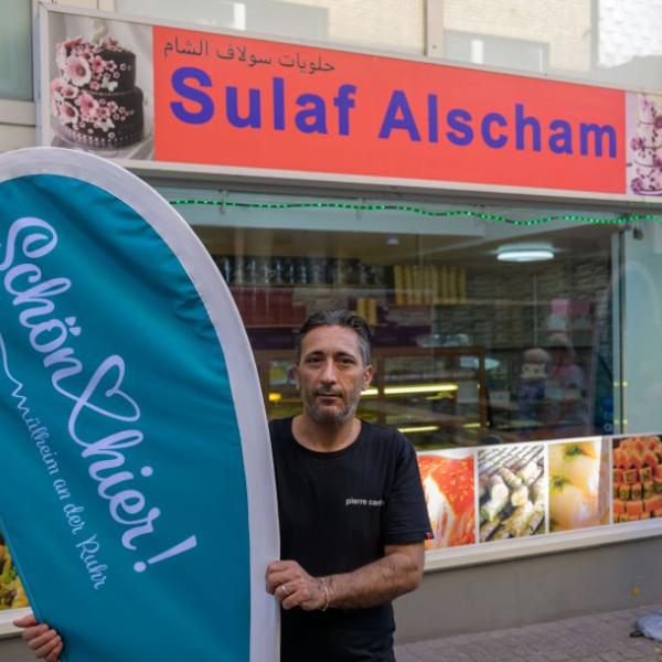 ein Mann steht vor seinem syrischen Süßigkeitenladen und hält eine hellblaue Schön hier!-Fahne fest | Foto: Walter Schernstein