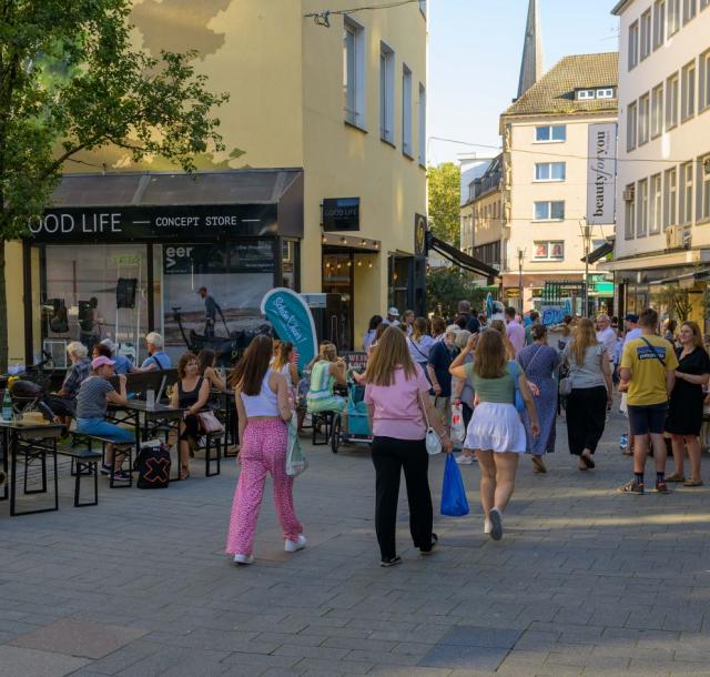 Auf dem Bild sind Menschen im Kohlenkamp in der Mülheimer Innenstadt zu sehen. Die Straße ist mit hellblauen Schön hier-Fahnen gesäumt. Es ist Sommer.